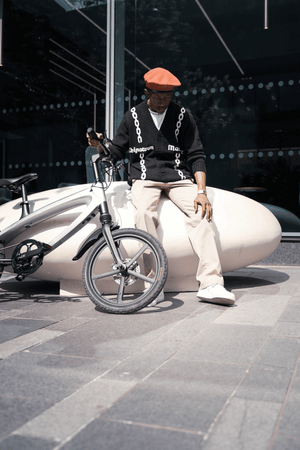 Stylish individual in a beret seated beside a grey e-bike on urban pavement, enjoying a moment of outdoor adventure.