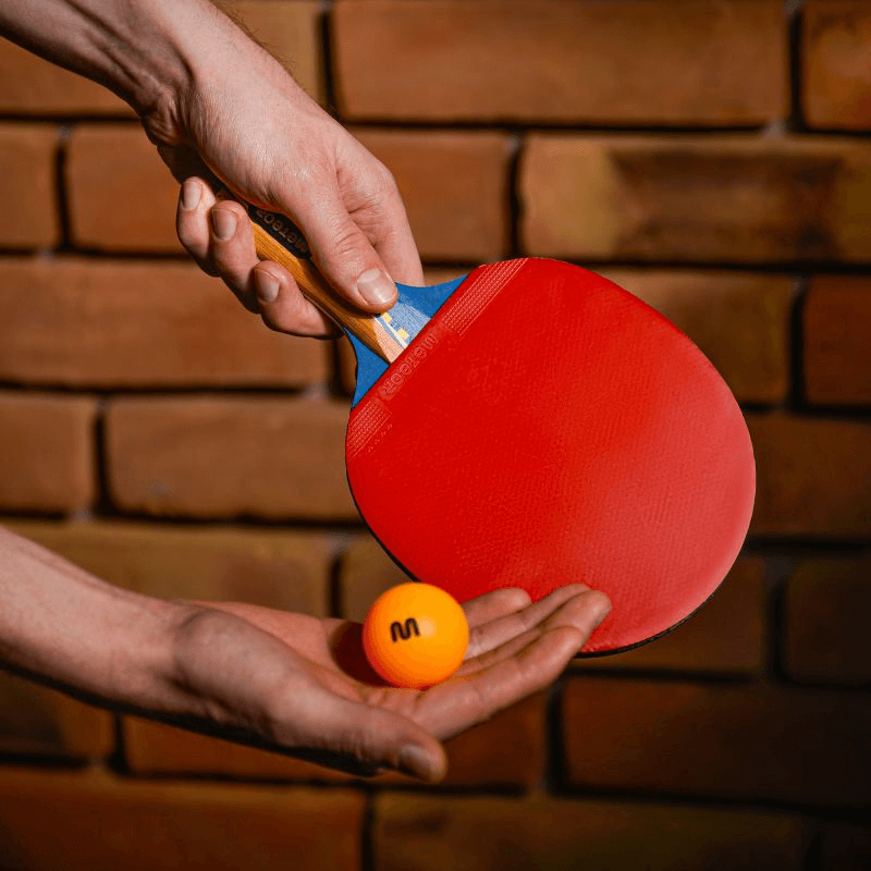 Hands holding a red Meteor Sirocco table tennis racket and a ping pong ball against a brick wall background.