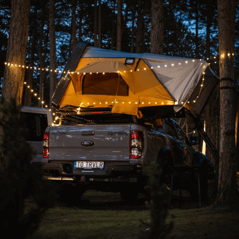 Offlander Fold 2 Sand Roof Tent mounted on a truck, illuminated by string lights in a scenic outdoor setting.