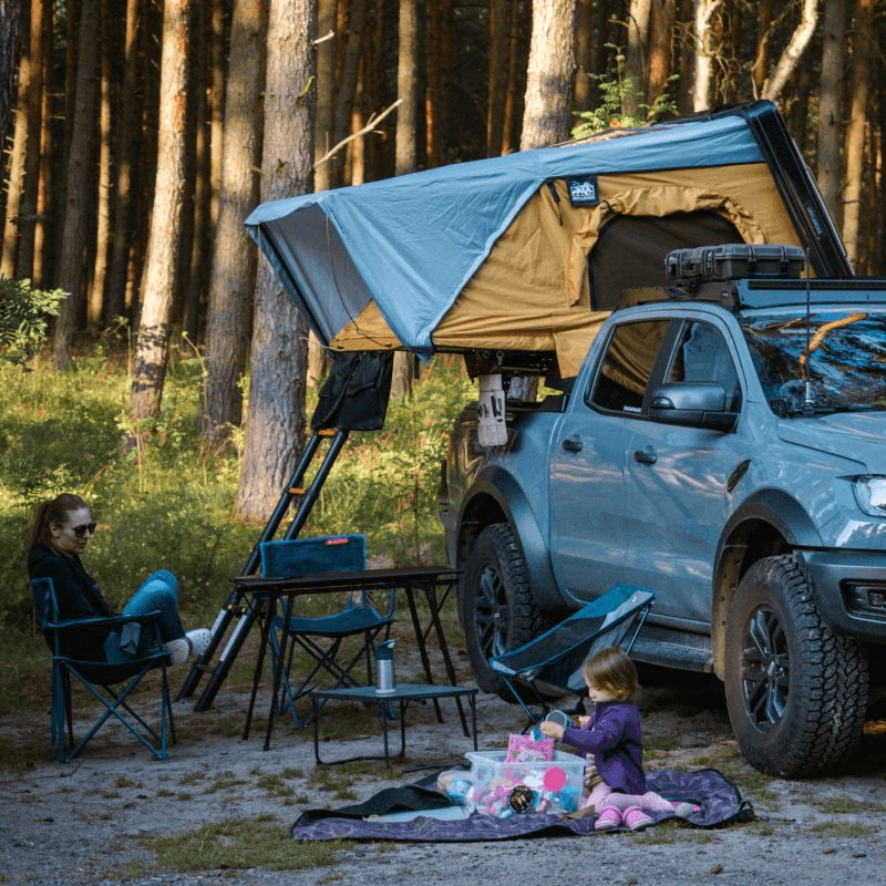 Family enjoying outdoor adventure with Offlander Fold 2 Sand Roof Tent mounted on vehicle in a forest setting.