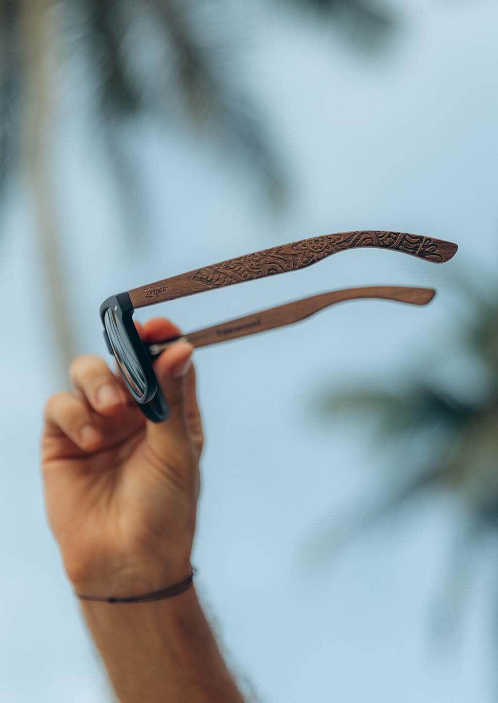 Engraved wooden sunglasses with mandala pattern held up outdoors, ideal for camping and adventures at revlando.com.